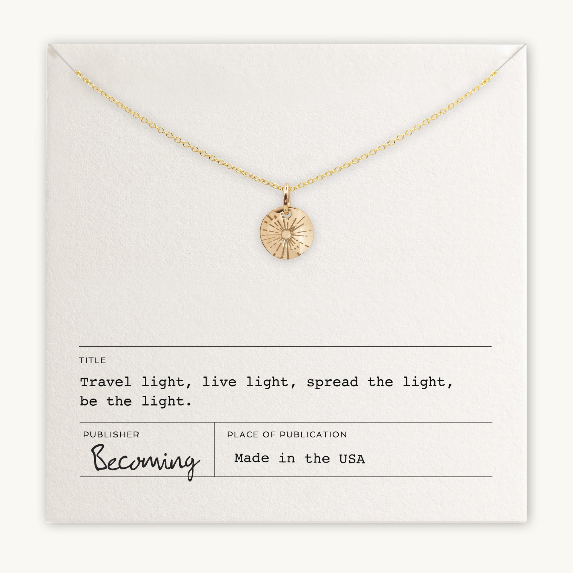 The Be The Light Necklace by Becoming Jewelry features a gold chain adorned with a radiant sun pendant and a small round charm, presented on a card inscribed with the message: &quot;Travel light, live light, spread the light, be the light.&quot; Manufactured in the USA.