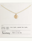 The Be The Light Necklace by Becoming Jewelry features a gold chain adorned with a radiant sun pendant and a small round charm, presented on a card inscribed with the message: "Travel light, live light, spread the light, be the light." Manufactured in the USA.