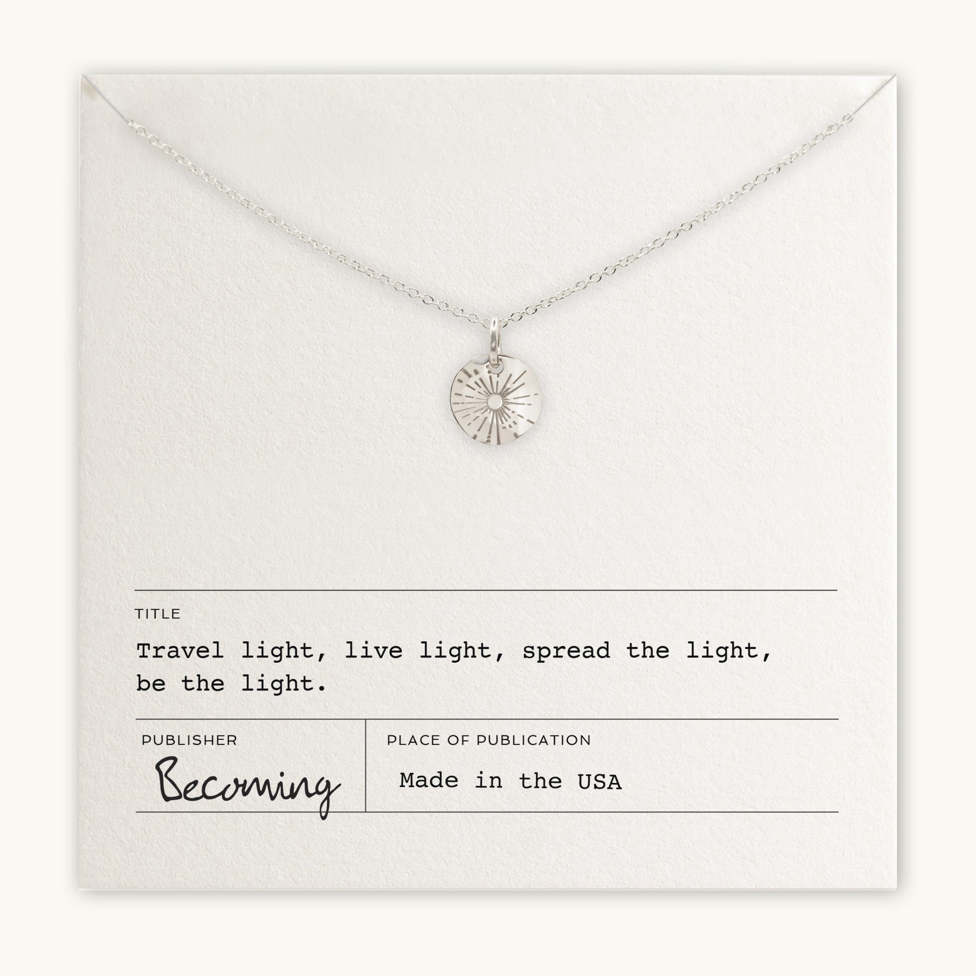 The "Be The Light Necklace" by Becoming Jewelry showcases a silver design with a sunburst pendant and a small round charm, elegantly presented on a card. The accompanying text states: "Travel light, live light, spread the light, be the light and shine brightly." Proudly made in the USA.