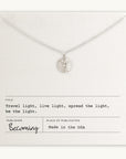 The "Be The Light Necklace" by Becoming Jewelry showcases a silver design with a sunburst pendant and a small round charm, elegantly presented on a card. The accompanying text states: "Travel light, live light, spread the light, be the light and shine brightly." Proudly made in the USA.