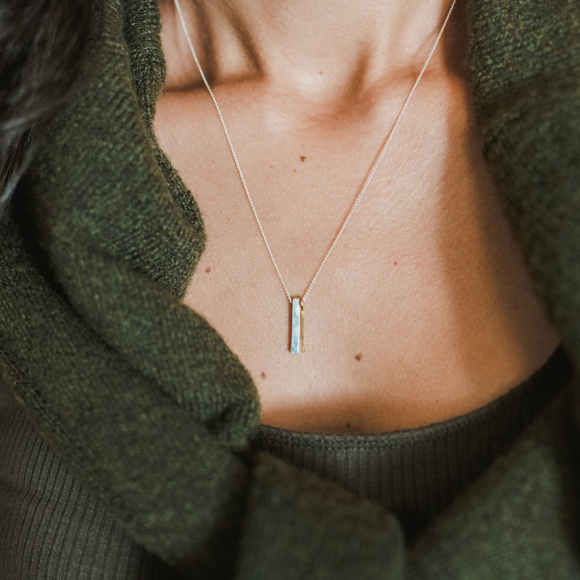 Close-up of a person wearing the Through Thick &amp; Thin Necklace by Becoming Jewelry. This best-selling, simple, thin necklace with a small vertical pendant makes the perfect gift. The person is dressed in a green sweater and a dark green top underneath.