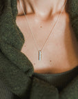 Close-up of a person wearing the Through Thick & Thin Necklace by Becoming Jewelry. This best-selling, simple, thin necklace with a small vertical pendant makes the perfect gift. The person is dressed in a green sweater and a dark green top underneath.