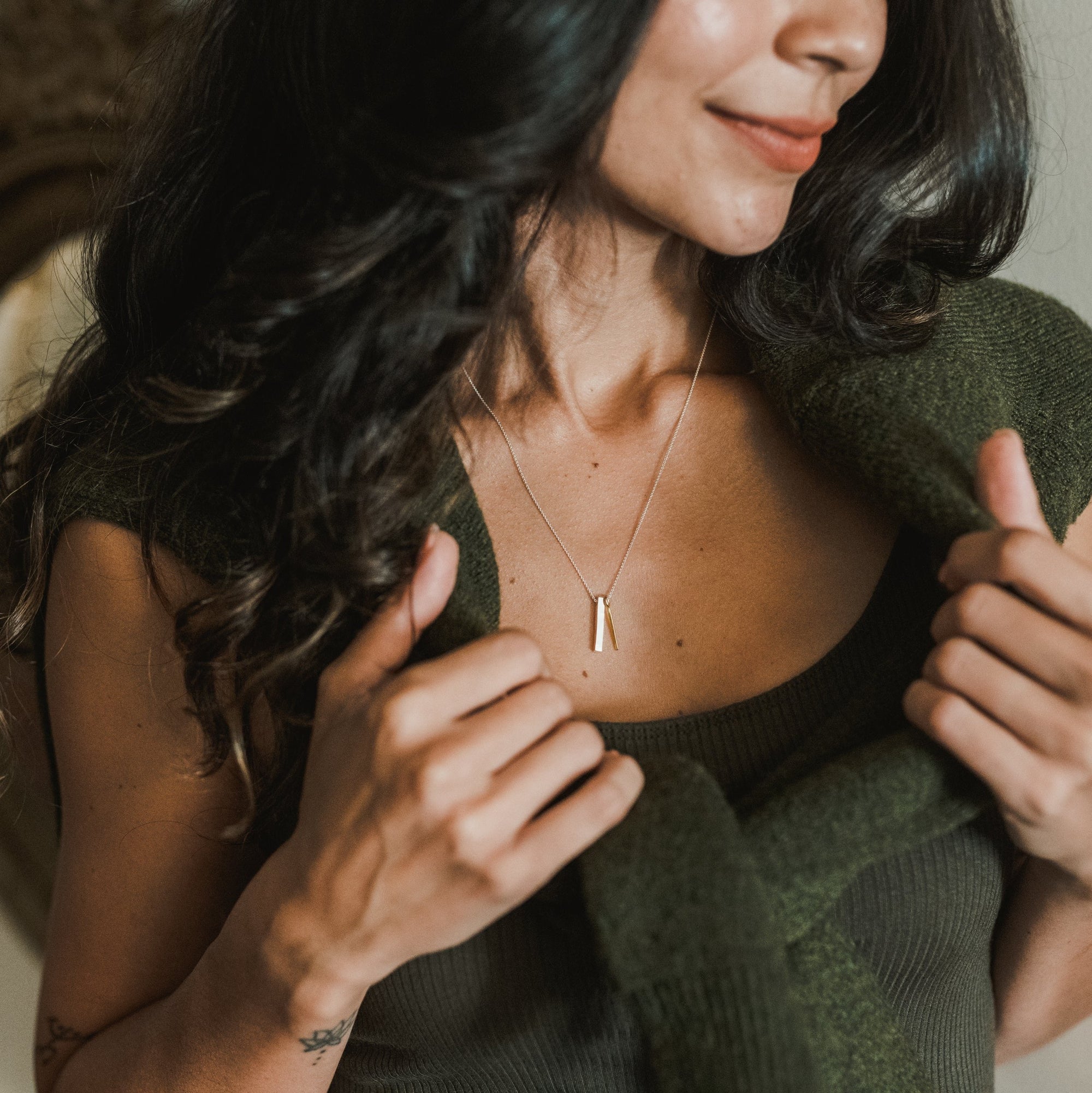 Close-up of a person with long, dark hair wearing a green top and Becoming Jewelry&#39;s best-selling Through Thick &amp; Thin Necklace, holding a green sweater draped over their shoulders.