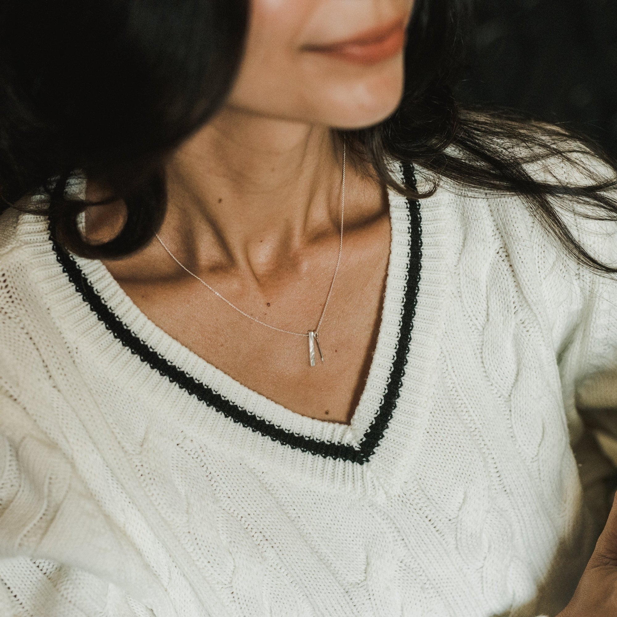 Close-up of a person wearing a white cable-knit sweater with a black-trimmed V-neck and the delicate silver &quot;Through Thick &amp; Thin Necklace&quot; by Becoming Jewelry. Their face is partially visible but out of focus, making it the perfect gift for someone special.