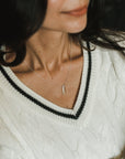 Close-up of a person wearing a white cable-knit sweater with a black-trimmed V-neck and the delicate silver "Through Thick & Thin Necklace" by Becoming Jewelry. Their face is partially visible but out of focus, making it the perfect gift for someone special.