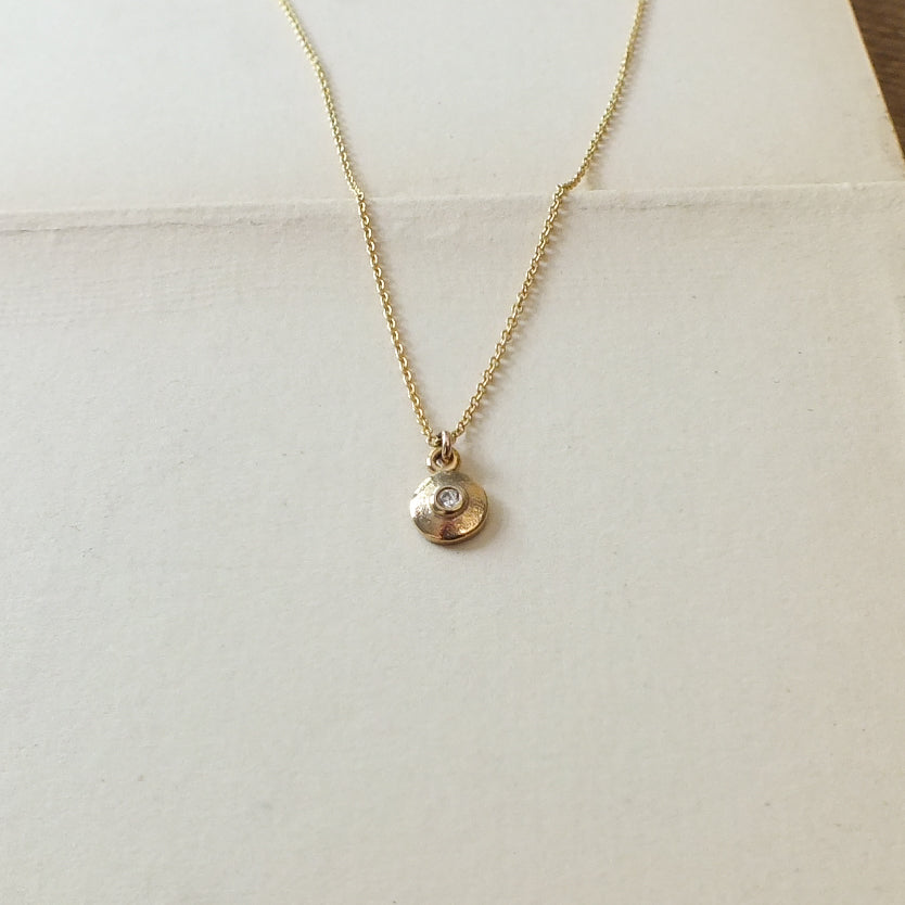 The Shine Necklace by Becoming Jewelry is a delicate gold accessory that features a small, round pendant adorned with a sparkling charm embedded with a single cubic zirconia, beautifully displayed on a light-colored surface.