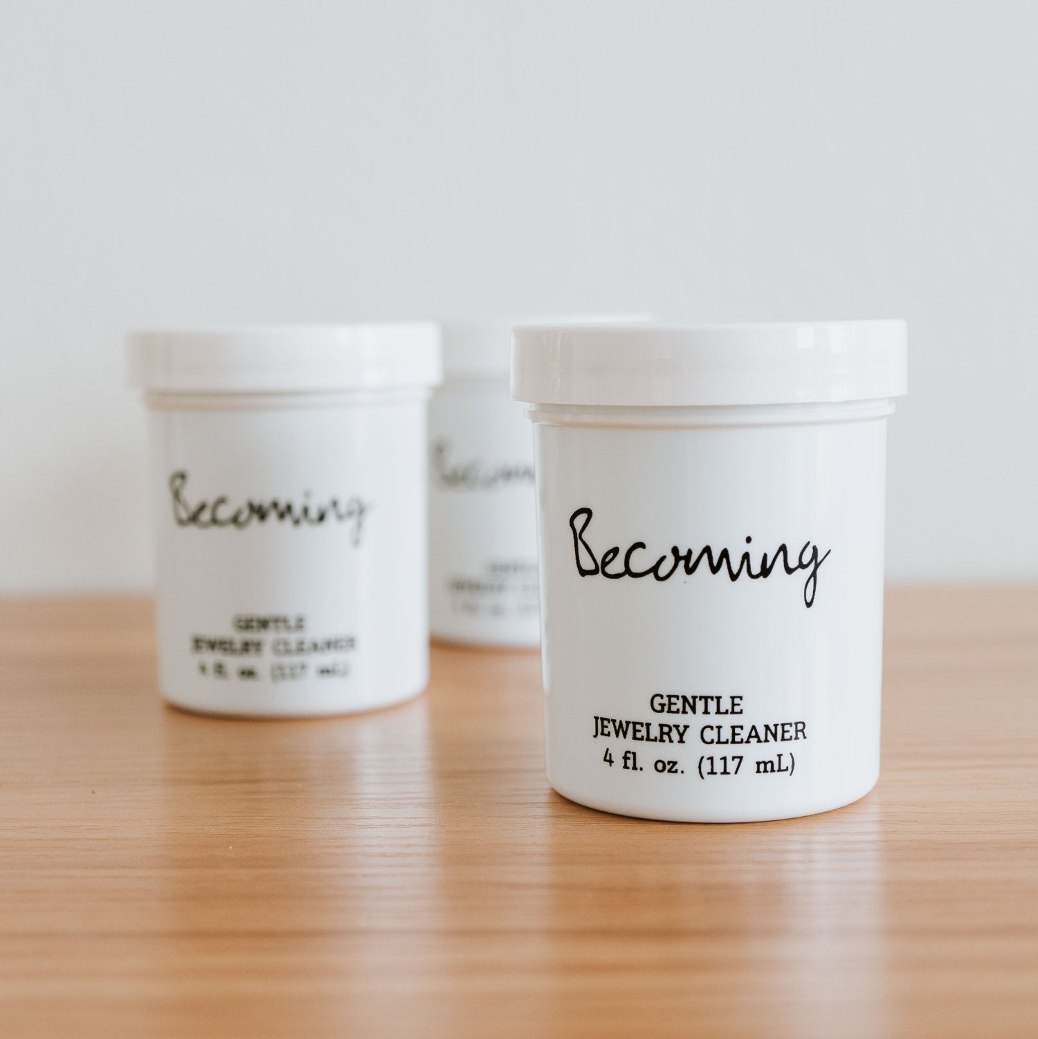 Three white containers labeled "Becoming Jewelry Cleaner" sit on a wooden surface. Inside, the liquid cleaner provides an ideal solution for your precious pieces, with each container equipped with a convenient dip tray to ensure every gem sparkles like new.