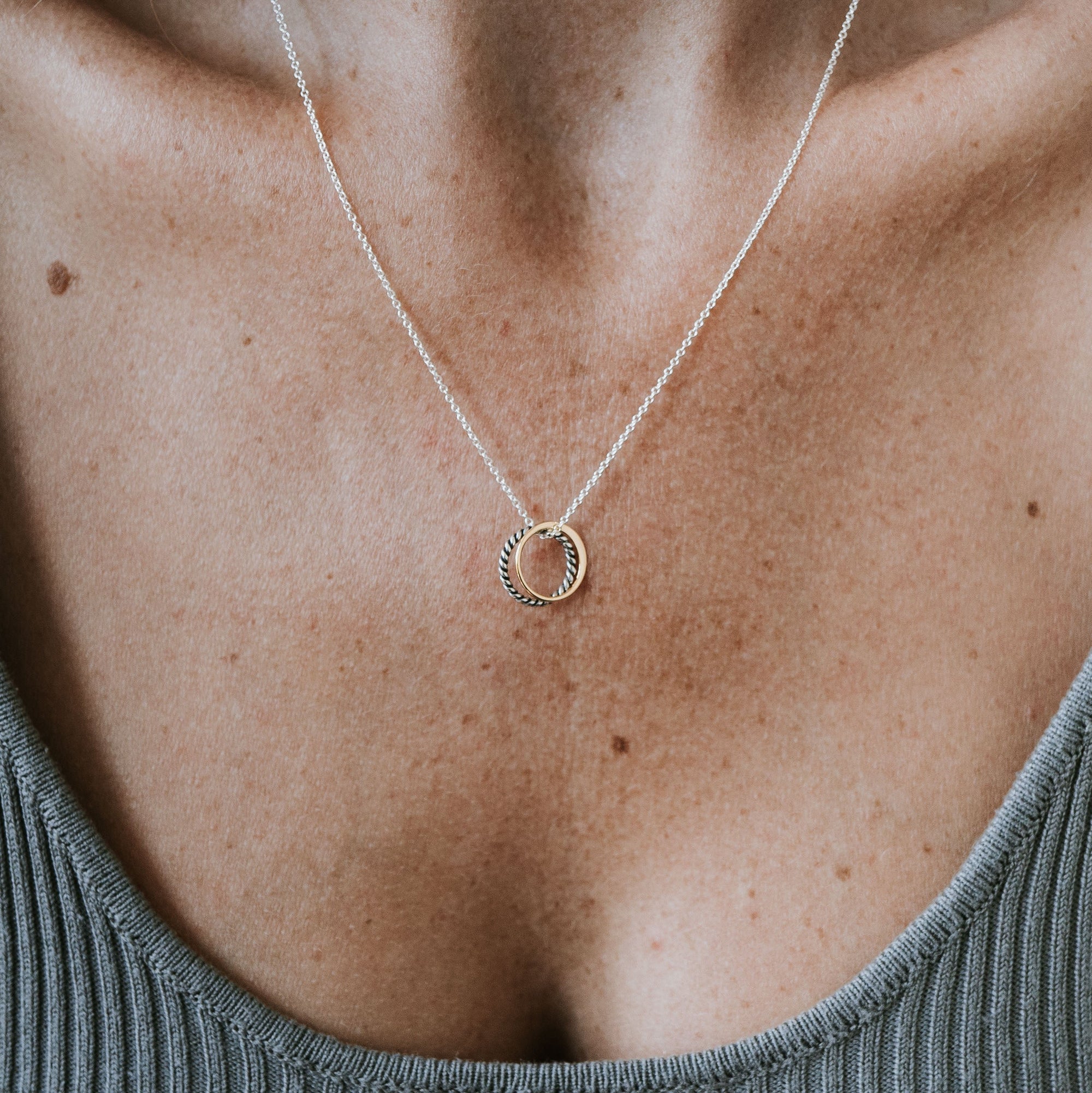 Close-up of a person&#39;s neck wearing the True Friends Necklace by Becoming Jewelry, featuring two mixed metal, textured circles as a pendant, shown against a ribbed grey top.